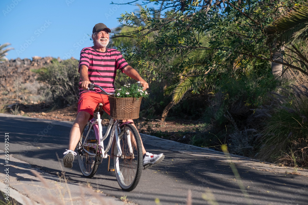 Happy senior man white hair and beard enjoying bicycle with fun. Colorful dressed, background of plants and palm trees. Blue sky on holiday
