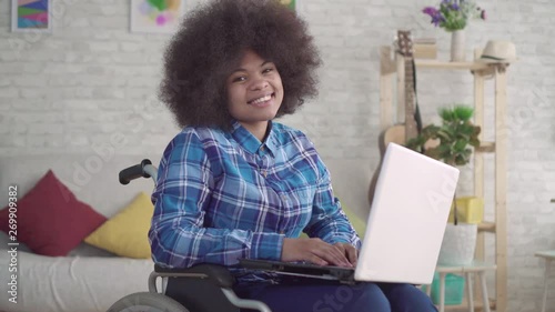 disabled african american woman with an afro hairstyle in a wheelchair uses a laptop looking at the camera photo