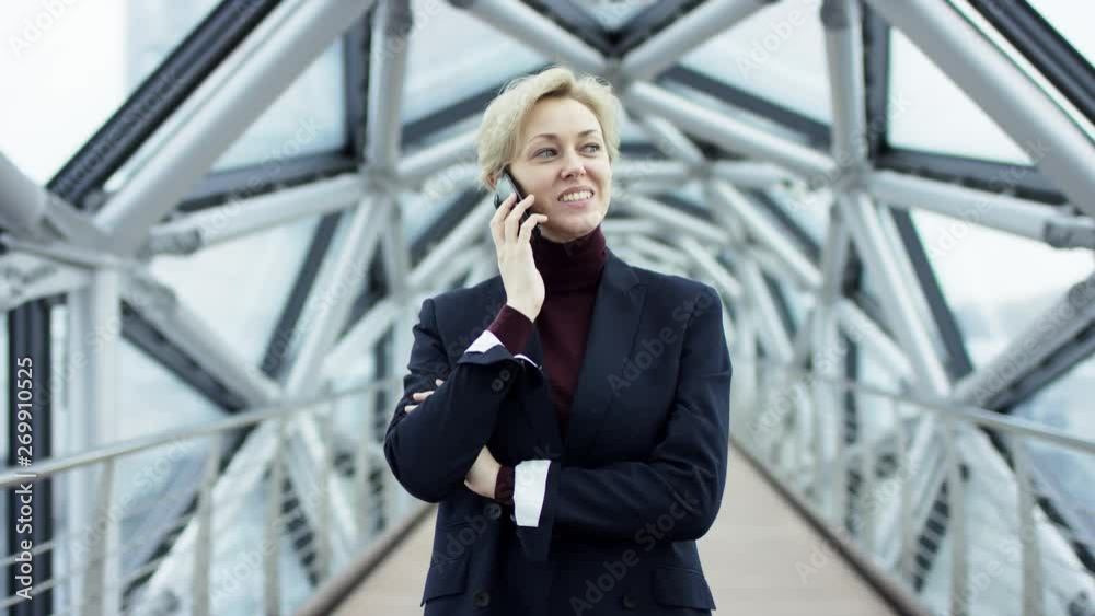 Portrait of a young beautiful business woman in a formal back suit, without glasses, walking around the city in the business center area, talking on the phone. She is discussing a business project Portrait of a young beautiful business woman in a formal back suit, without glasses, walking around the city in the business center area, talking on the phone. She is discussing a business project