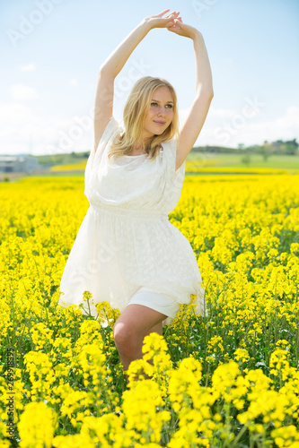Happy girl stretch oneself in rape seed flowers field posing in white dress