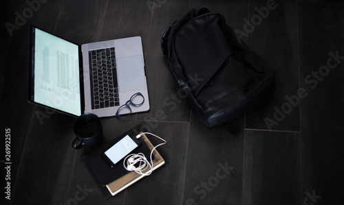 Top view of black backpack near laptop, smartphone with earphones, cup of coffee, notebook and glasses on the floor