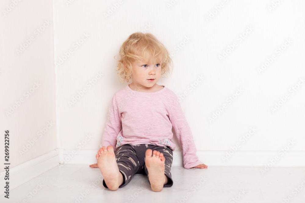 Naklejka premium curly blonde baby girl in comfortable home casual clothes is lying on the floor in a bright white room smiling and looking at the camera