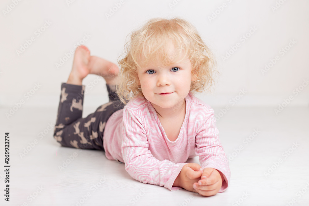 curly blonde baby girl in comfortable home casual clothes is lying on the floor in a bright white room smiling and looking at the camera curly blonde baby girl in comfortable home casual clothes is lying on the floor in a bright white room smiling and looking at the camera