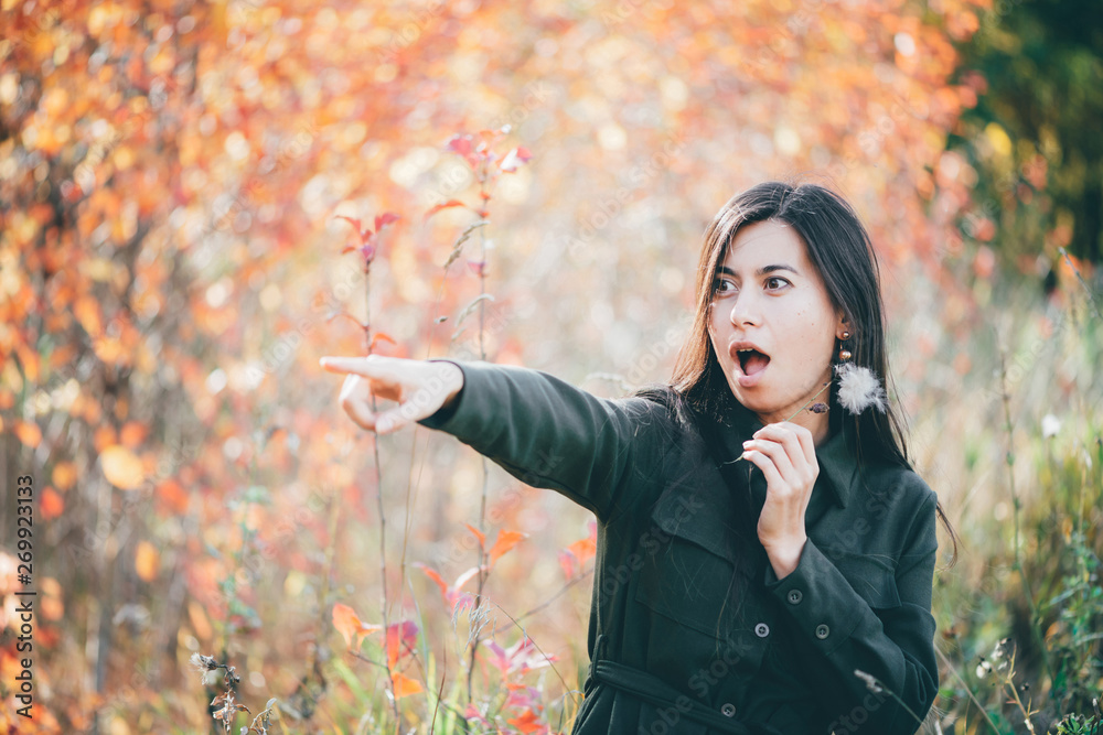 Shocked girl with surprised look points finger away. Beautiful girl with long natural black hair in shock condition on bokeh background with autumn leaves. Female beauty portrait among vivid foliage. Shocked girl with surprised look points finger away. Beautiful girl with long natural black hair in shock condition on bokeh background with autumn leaves. Female beauty portrait among vivid foliage.