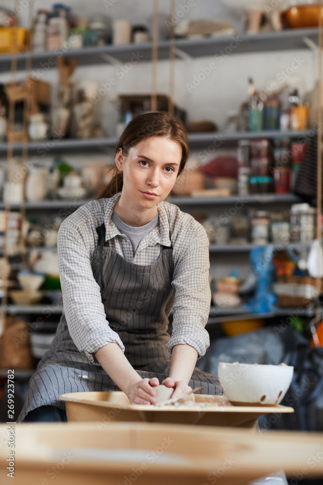 Serious displeased young woman with ponytail forming vase on pottery wheel and looking at camera in workshop