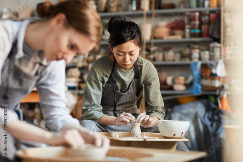 Fototapeta Serious skilled young Asian female potter in apron sitting at pottery wheel and