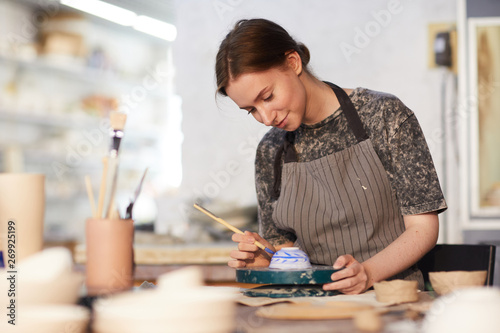 Content attractive skilled young lady in apron standing at table and drawing blue leaves on ceramic bowl in pottery workshop