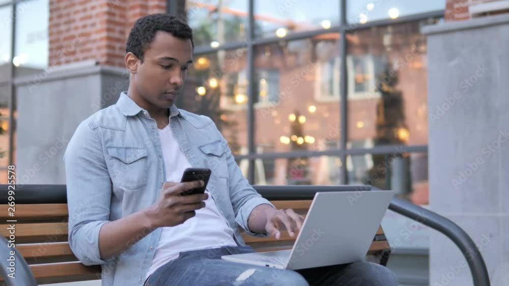 African Man Using Smartphone and Laptop, Sitting on Bench African Man Using Smartphone and Laptop, Sitting on Bench