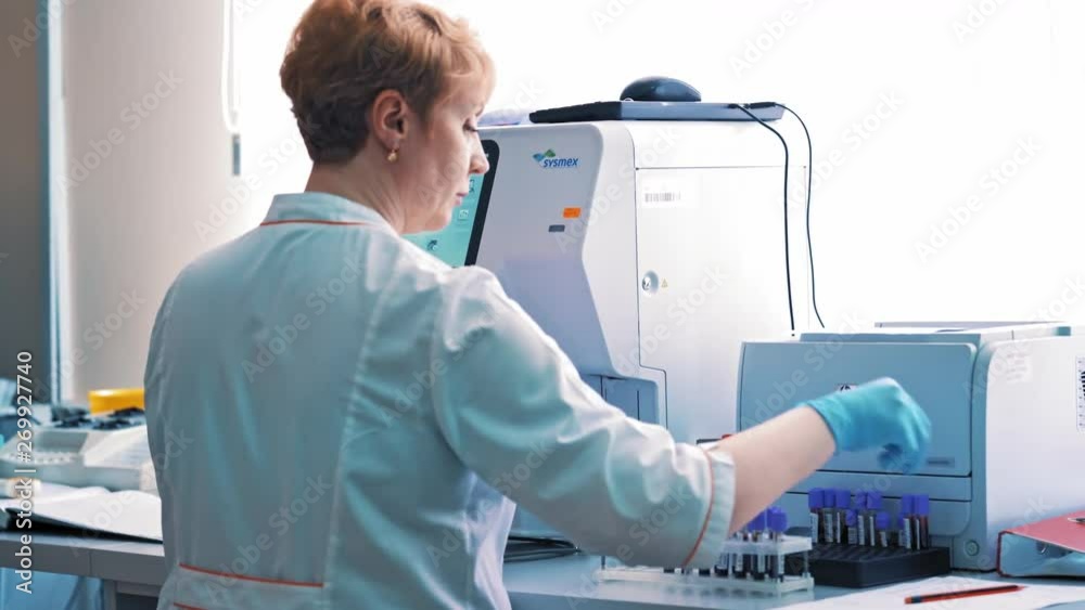 Woman worker in medical gown holds the vial close to the medical aparatus in the laboratory. Laboratory technician works with test tubes and writes the results on paper.