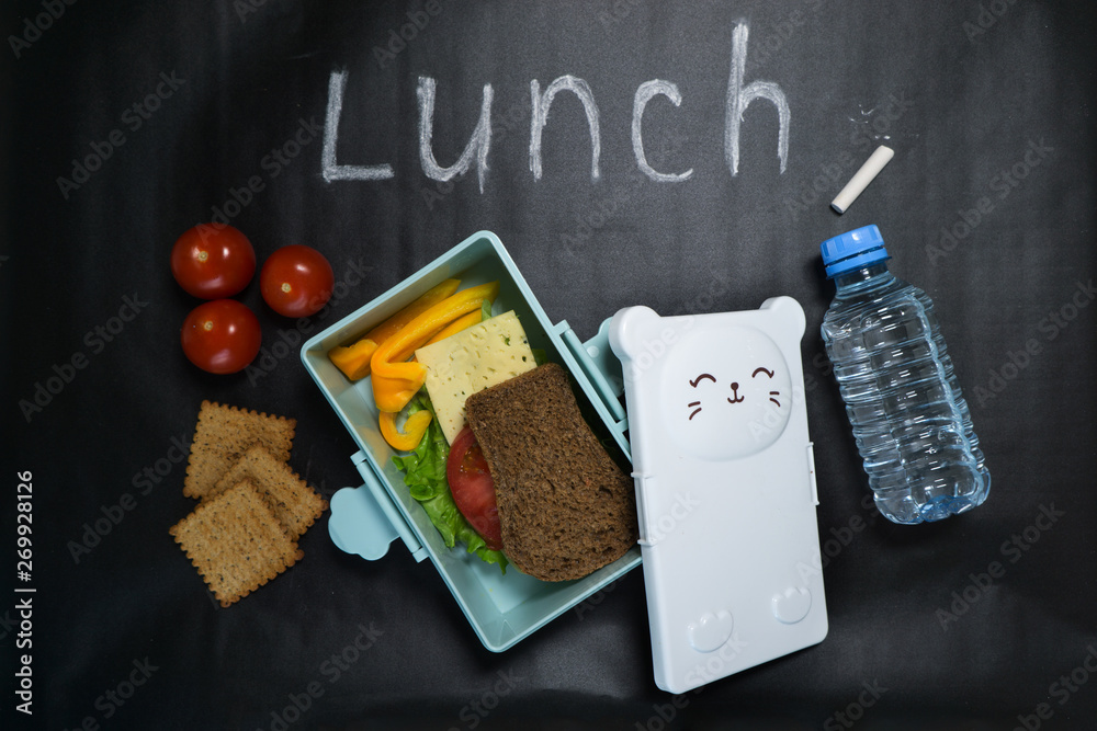 Obraz premium open lunch box with a sandwich of whole grain bread, cheese, green salad, tomato, cucumber and a bottle of water on a black background with colored crayons and lunch inscription written in white chalk