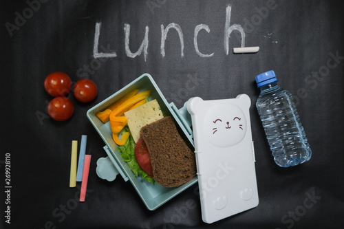 open lunch box with a sandwich of whole grain bread, cheese, green salad, tomato, cucumber and a bottle of water on a black background with colored crayons and lunch inscription written in white chalk