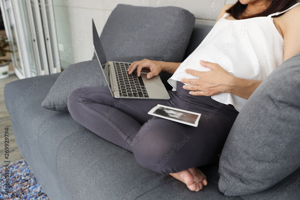 portrait of beautiful asian pregnant woman using computer and listening to music while looking ultrasound photo on sofa in living room portrait of beautiful asian pregnant woman using computer and listening to music while looking ultrasound photo on sofa in living room