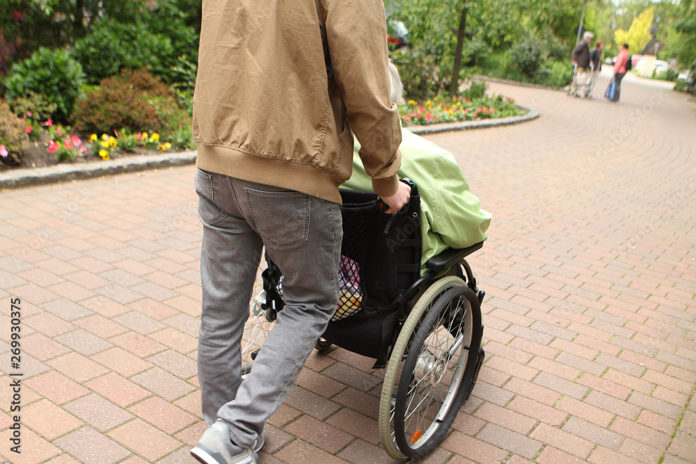 Obraz premium A man in gray jeans carries an elderly woman, a disabled person in a wheelchair on the street for a walk, rear view