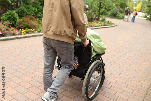 A man in gray jeans carries an elderly woman, a disabled person in a wheelchair on the street for a walk, rear view