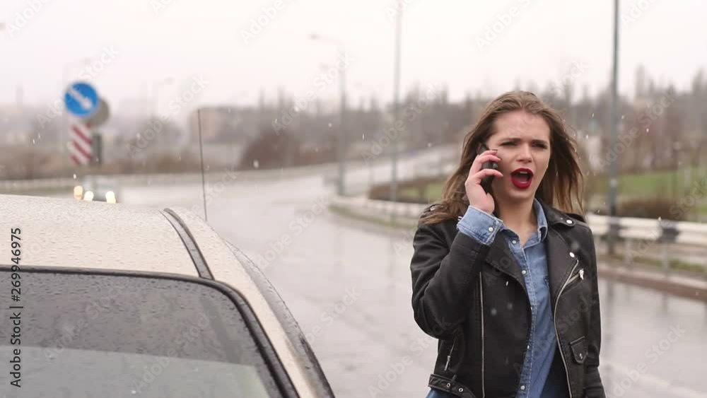 A young girl is standing in the rain with snow on an empty road outside the city with a phone, her car is broken and she calls and asks for help. A young girl is standing in the rain with snow on an empty road outside the city with a phone, her car is broken and she calls and asks for help.