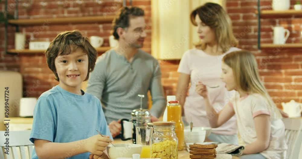 Portrait of the cute little Caucasian boy smiling to the camera in the kitchen while his family having breakfast on the background. Portrait of the cute little Caucasian boy smiling to the camera in the kitchen while his family having breakfast on the background.