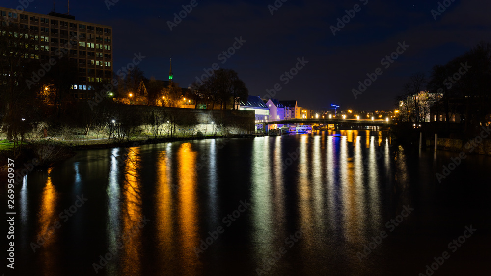 The German city of Kassel, which lies at the river Fulda, at night. Excursion ships are located on the jetty. The lights of the city are reflected in the water.
