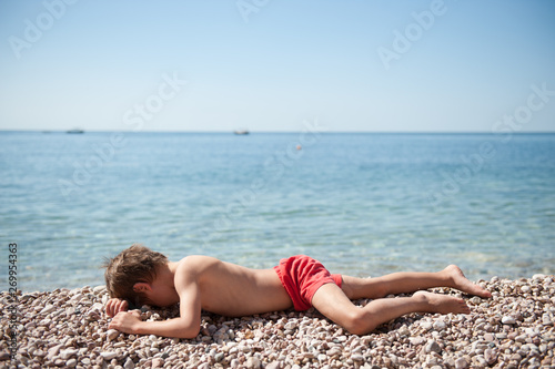 tired little boy in red shorts lying on stone beach near ocean sunbathe during summer holiday travel activity with copy space