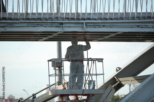 Professional worker in a protective suit and mask paints a bridge from the spray gun stand on the hydraulic lift.