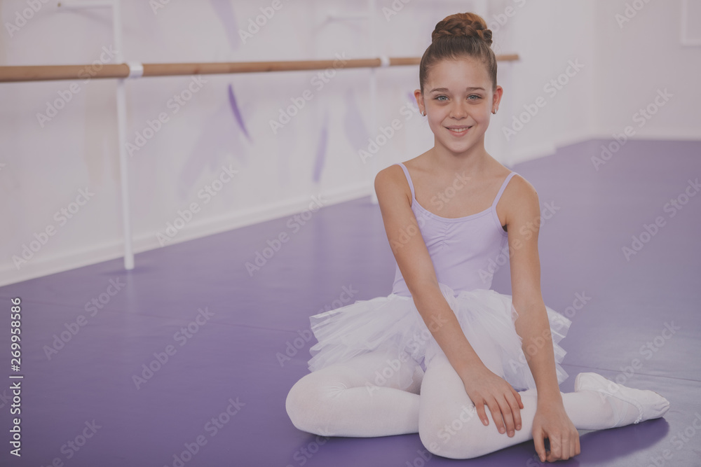 Happy beautiful young ballerina girl smiling to the camera, sitting on the floor at ballet dance school. Charming little ballet dancer resting after exercising,copy space Happy beautiful young ballerina girl smiling to the camera, sitting on the floor at ballet dance school. Charming little ballet dancer resting after exercising,copy space
