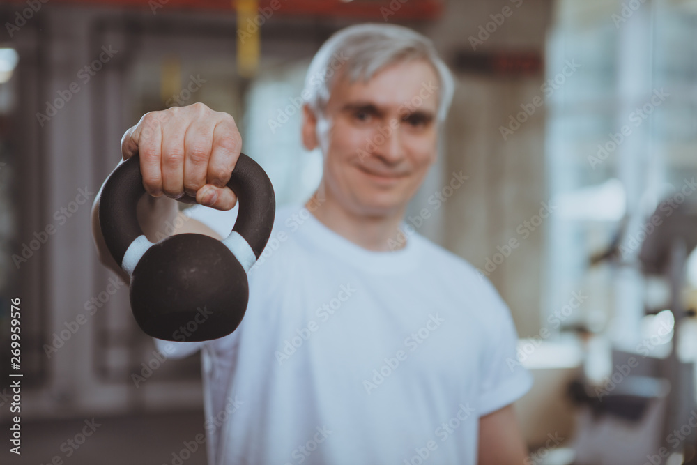 Selective focus on a kettlebell senior happy sportsman is holding out to  the camera. Cheerful elderly man enjoying working out at sports studio,  lifting weights, copy space. Health, strength concept Stock Photo