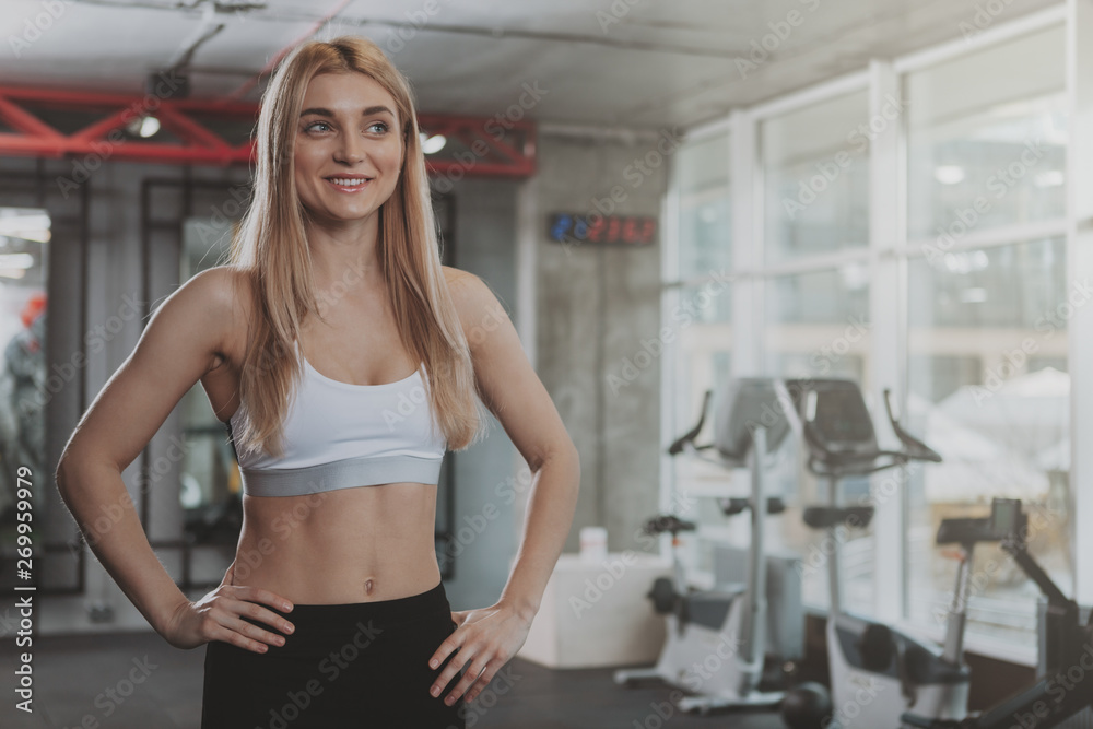 Charming blond haired young fitness woman in sports top and leggings posing confidently after working out. Cheerful female athlete enjoying morning workout at sport studio, copy space