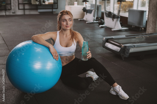 Top view shot of a beautiful healthy sportswoman leaning on a fitball, sitting on the gym floor with a bottle of water. Attractive female athlete relaxing after exercising on fitness ball, copy space