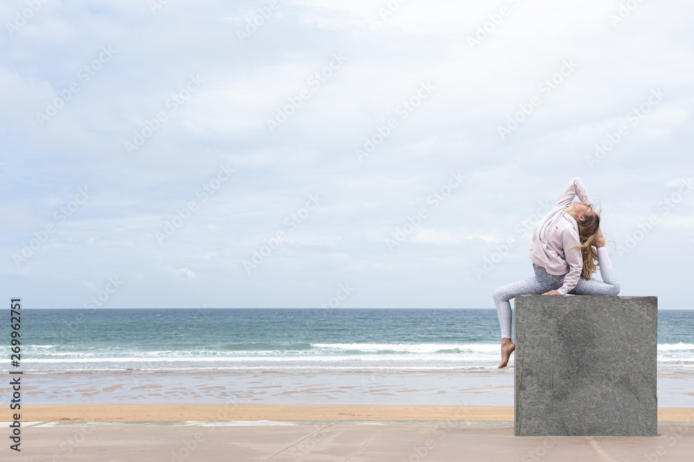 Woman yogi doing the pigeon pose on a stone cube on the beach with the sea in the background