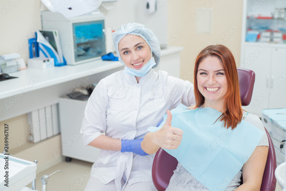 Obraz premium A female dentist and a red-haired patient in an office after dental treatment are looking at the camera with happy faces. Happy woman with her doctor after taking shows sign for shelter.