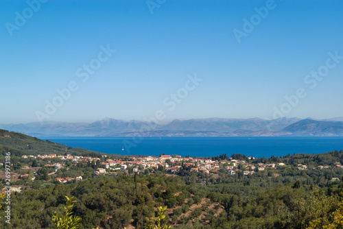 Beautiful panoramic view of the Ionian island of Corfu with olive trees, a small village, the blue waters of the Ionian Sea and high mountains in the background. Half landscape, half blue sky.