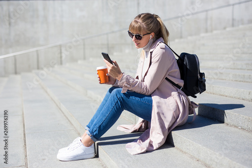 lifestyle and travel concept - outdoor portrait of young woman sitting on stairs with smartphone and coffee