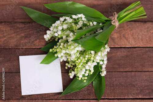 bouquet of delicate white lilies of the valley on a brown wooden background with a white paper sheet for text