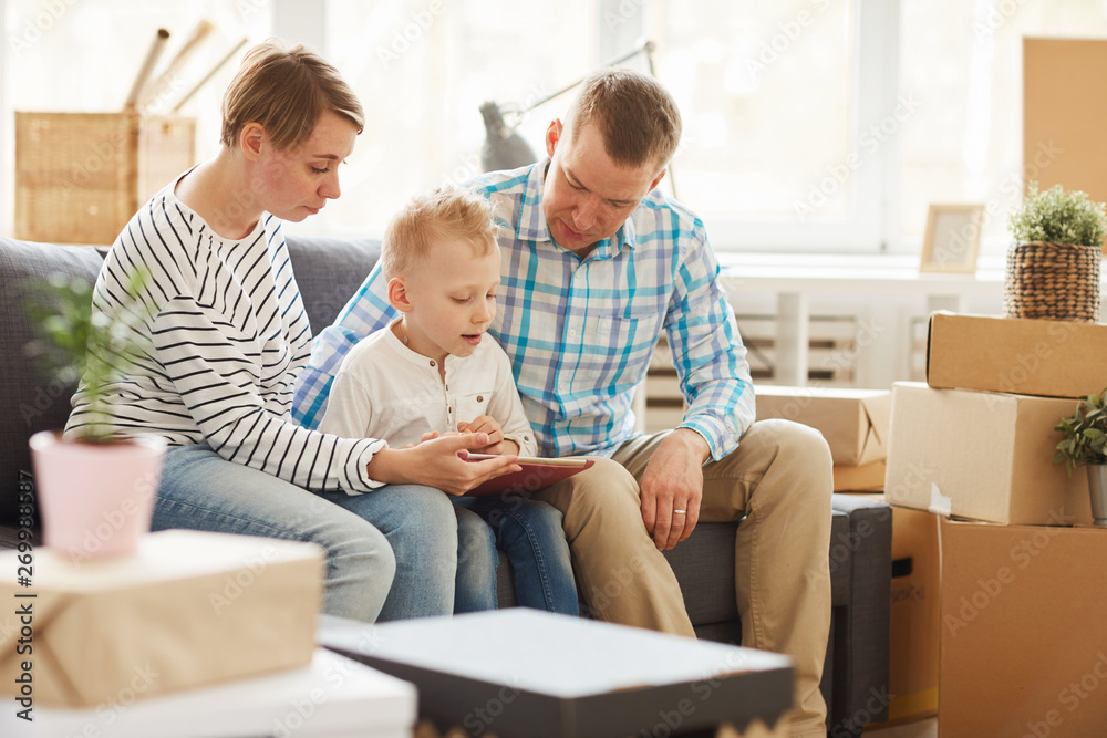 Content curious blond-haired son sitting with parents on sofa in living room with stacks of boxes and reading book on tablet while doing home task. Content curious blond-haired son sitting with parents on sofa in living room with stacks of boxes and reading book on tablet while doing home task.