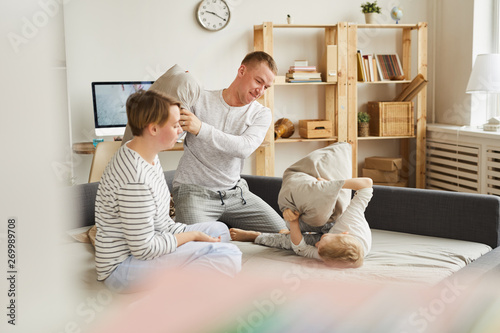 Cheerful excited active young parents in pajamas sitting on sofa bed and playing pillow fight with son