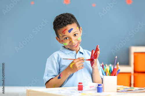 Fototapeta Naklejka Na Ścianę i Meble -  Cute African-American boy with hands and face in paint at home