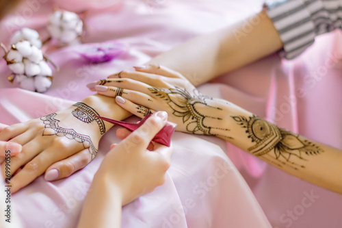 Female wrists painted with traditional oriental mehndi ornaments. Process of painting womens hands with henna, preparing for indian wedding. Pink fabric with folds, flowers and candles on background.