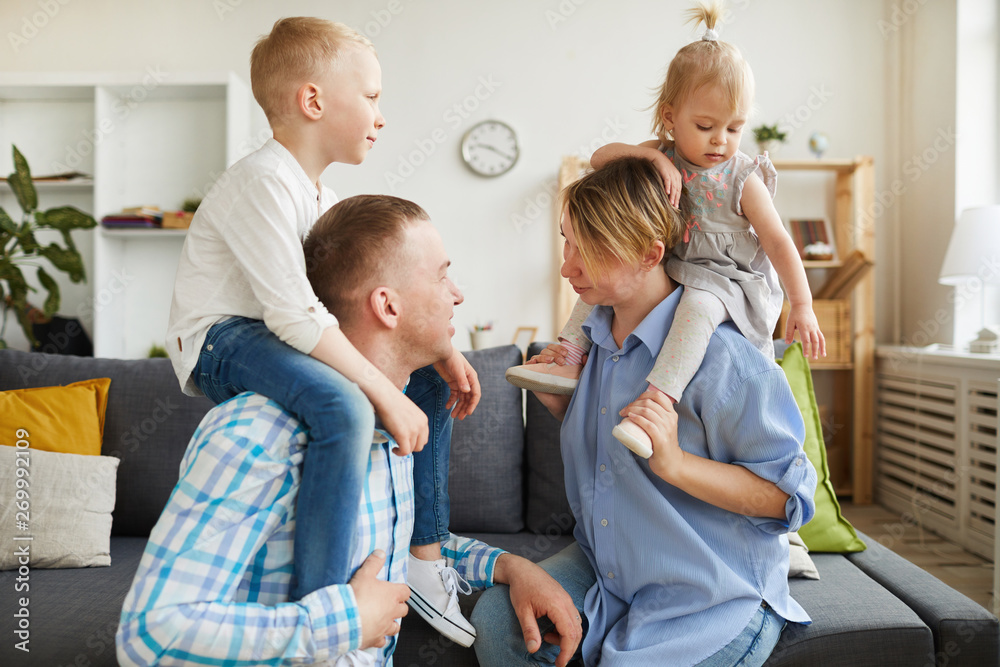Happy young parents riding children on necks while playing with them at home, loving parents chatting each other Happy young parents riding children on necks while playing with them at home, loving parents chatting each other