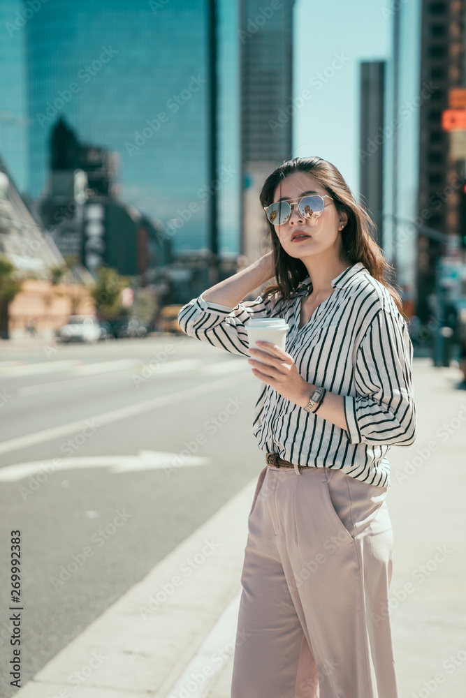 Fashion asian young girl ceo holding paper cup of coffee in hands standing on street. female worker waiting for transport and taxi in morning going work. woman in business area enjoy sunshine outdoor