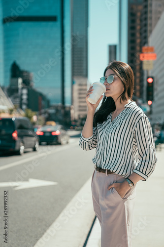 Professional young urban casual business woman in los angeles city out of concert hall standing on street having hot cup coffee waiting for yellow cab taxi. asian office lady drinking tea for morning