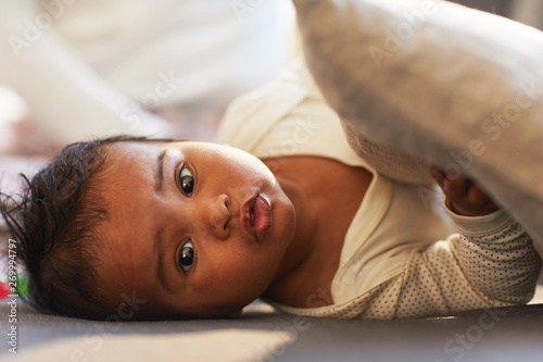 Portrait of adorable African baby boy with puffy lips lying on comfortable sofa in living room and holding pillow