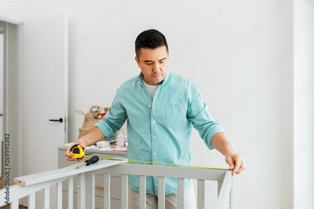 parenthood, fatherhood and nursery concept - middle-aged father with ruler measuring baby bed at home