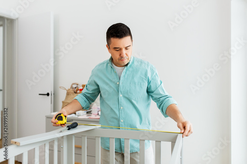 parenthood, fatherhood and nursery concept - middle-aged father with ruler measuring baby bed at home