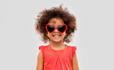 childhood, valentine's day and summer concept - happy little african american girl in heart shaped sunglasses over grey background