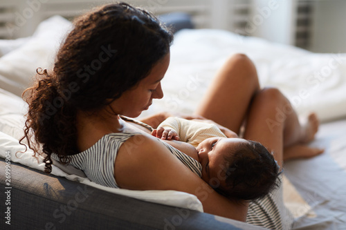 Peaceful loving young African mother sitting on bed and leaning on headboard while feeding baby with breast