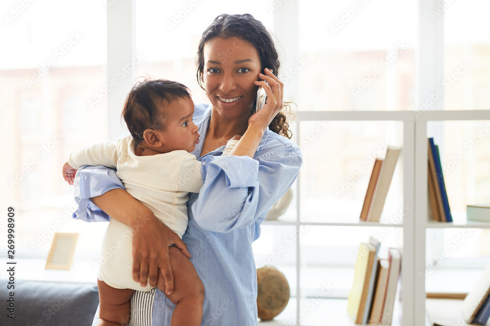 Positive confident modern young African mom standing against shelves and holding baby in arm while talking on mobile phone and looking at camera Positive confident modern young African mom standing against shelves and holding baby in arm while talking on mobile phone and looking at camera