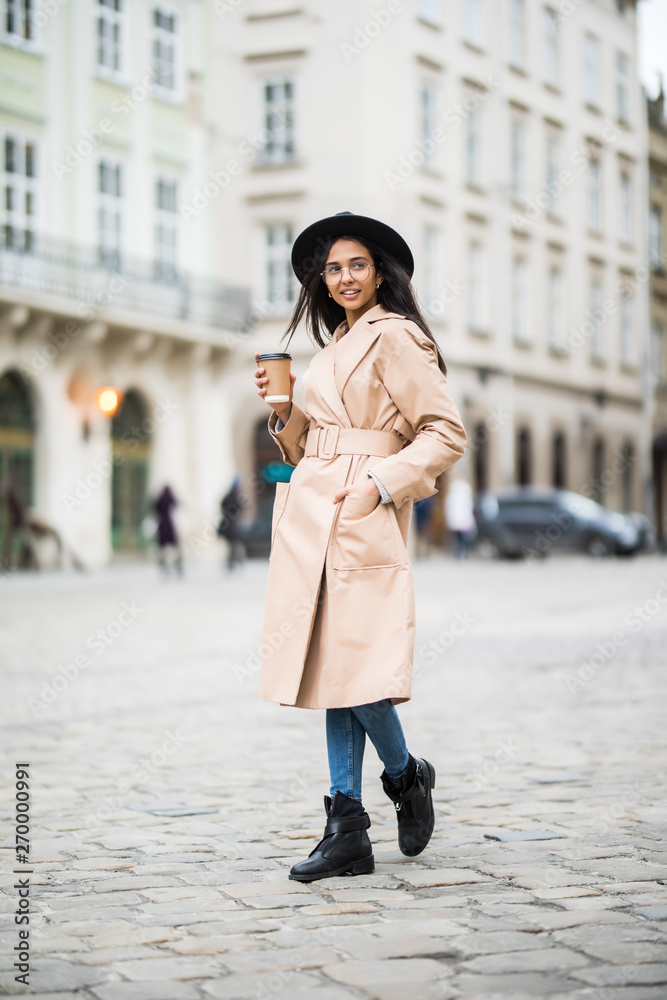 Young woman wearing coat and black hat walking in the autumn city street and drinking take away coffee in paper cup. Breakfast to go.