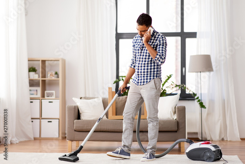 household and cleaning concept - indian man with vacuum cleaner calling on smartphone at home
