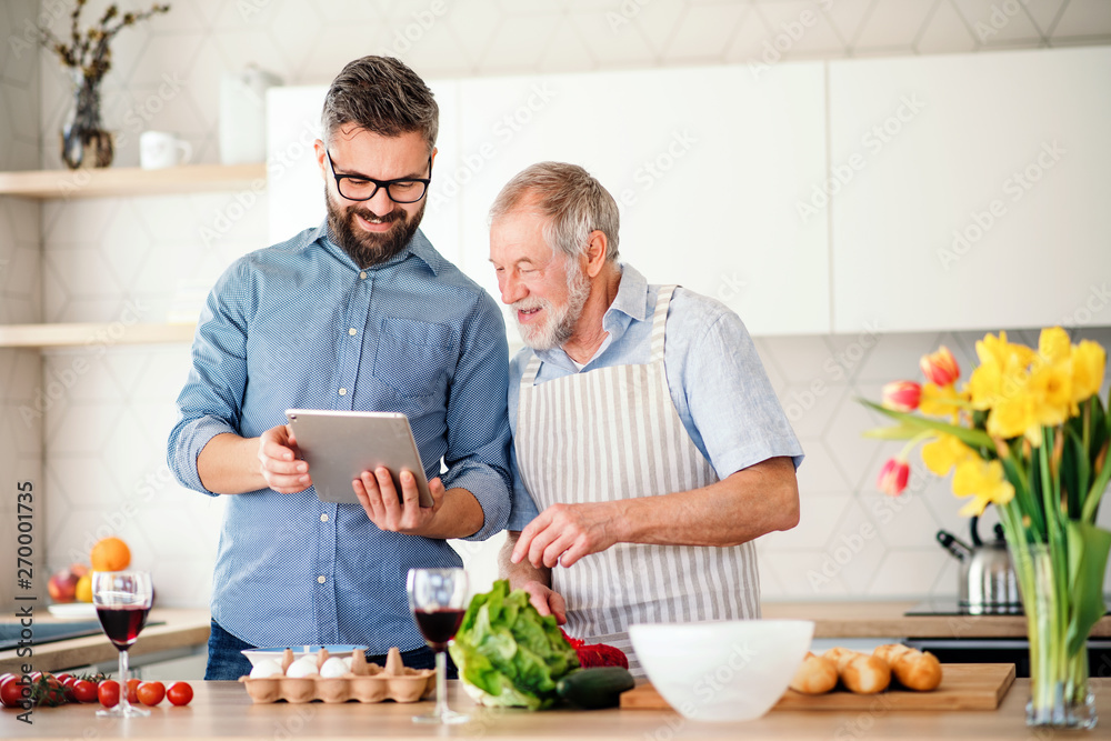 Adult hipster son and senior father indoors in kitchen at home, using tablet. Adult hipster son and senior father indoors in kitchen at home, using tablet.