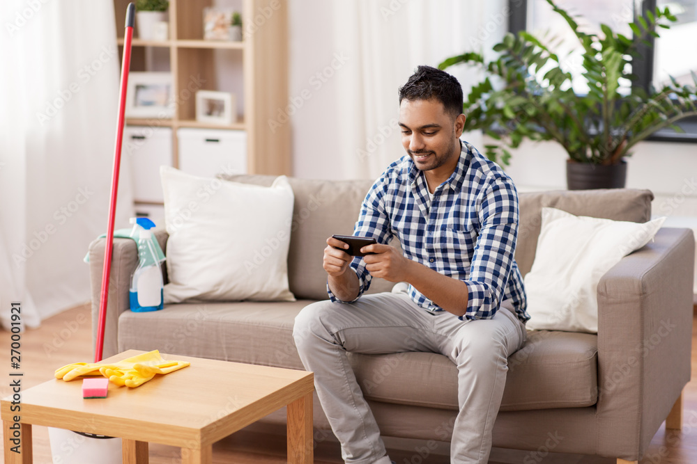 household and technology concept - indian man playing game on smartphone after cleaning home