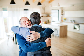 An adult hipster son and senior father indoors at home, hugging.